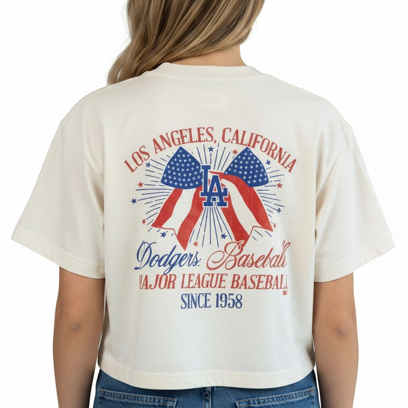 Load image into Gallery viewer, Back view of a woman wearing a relaxed fit '47 Brand Los Angeles Dodgers crop top featuring a patriotic red, white, and blue USA bow graphic, fireworks, and vintage baseball-inspired artwork celebrating Los Angeles, California.
