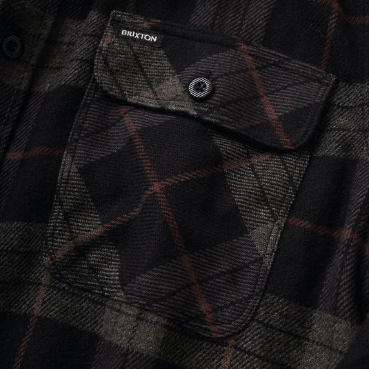 Close-up of a dark plaid flannel shirt pocket with a snap flap. The fabric is black and charcoal gray with thin brown lines, and the Brixton brand label is visible near the top.