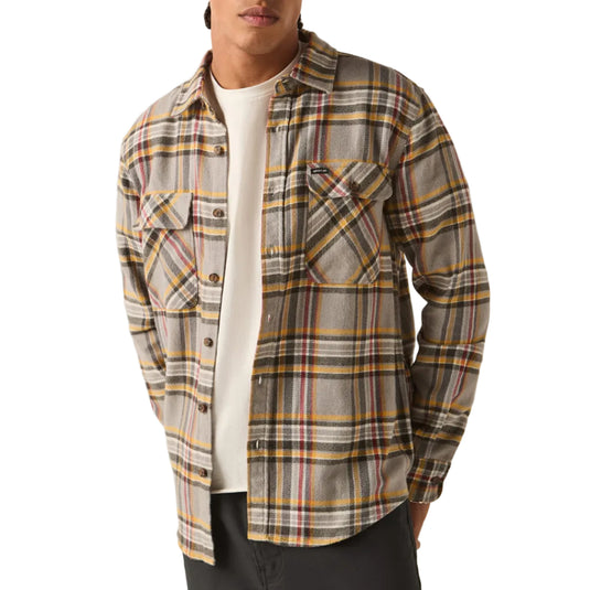 A man wearing a beige and yellow plaid button-up shirt over a white t-shirt, paired with a red knit beanie and dark gray pants, standing against a white background.