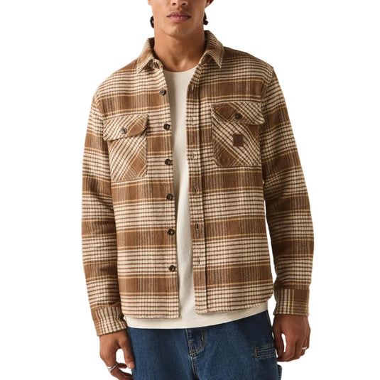 A model wearing a brown and beige plaid button-up overshirt over a white t-shirt, blue jeans, and a red knit beanie, standing against a plain off-white background.