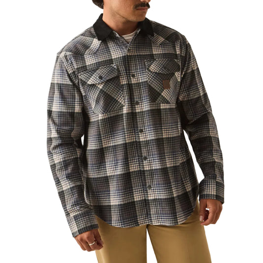 A man wearing a black and gray plaid button-up flannel shirt, tan pants, and a black baseball cap with a logo patch, standing against a plain white background.