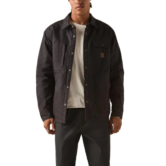 A man modeling a dark charcoal work jacket over a white t-shirt, paired with dark gray pants, black shoes, and a red beanie, standing against a plain off-white studio background.