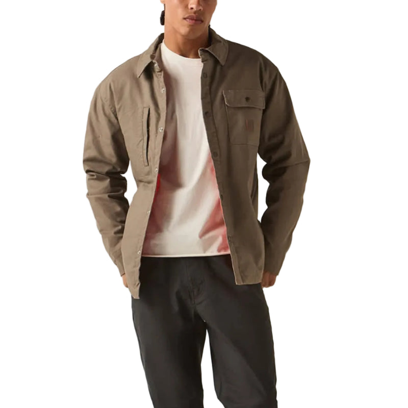 Load image into Gallery viewer, A man stands against a plain light beige studio background wearing a taupe-brown overshirt over a white graphic T-shirt, dark charcoal pants, black shoes, and a white baseball cap with a red brim and a circular patch.
