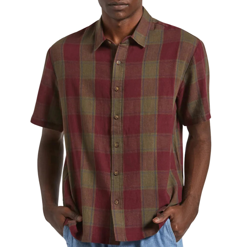 Load image into Gallery viewer, A man wearing a short-sleeve button-up plaid shirt in red and olive green, paired with light blue jeans, against a plain light background.
