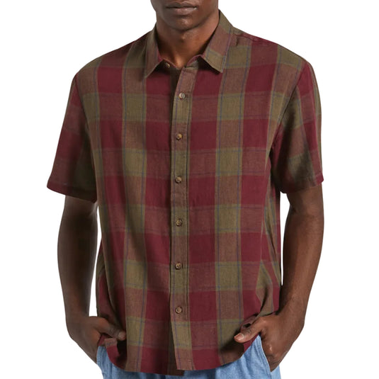 A man wearing a short-sleeve button-up plaid shirt in red and olive green, paired with light blue jeans, against a plain light background.