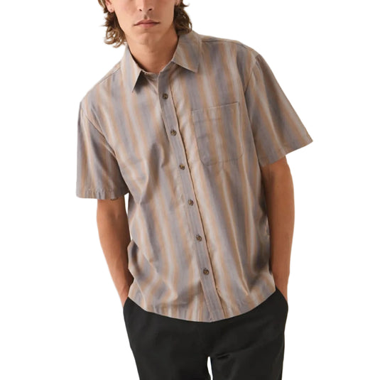 A man wearing a short-sleeve button-up shirt with vertical stripes in beige, brown, and muted blue, paired with black pants, standing against a plain off-white studio background.