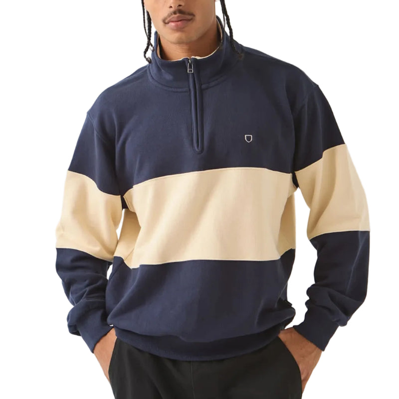 Load image into Gallery viewer, A man wearing a navy and beige color-blocked quarter-zip sweatshirt, black pants, and a dark flat cap, standing against a light gray studio background.

