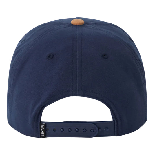 A navy blue snapback cap shown from the back, featuring a small tan button on top and an adjustable dark strap with a small black brand tag reading 'BRIXTON'.