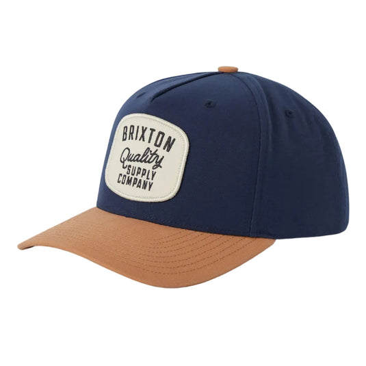 A navy blue baseball cap with a tan visor and a white Brixton patch on the front, photographed on a light gray background.