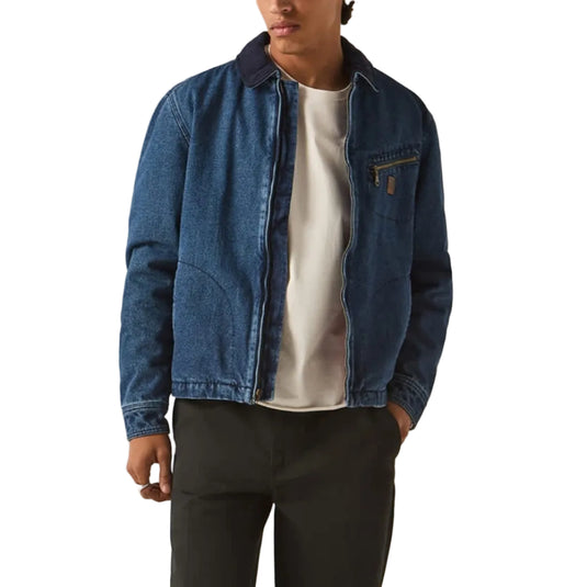 A man modeling a blue denim jacket over a white t-shirt, paired with black trousers and a dark gray newsboy cap, standing against a plain light beige studio background.