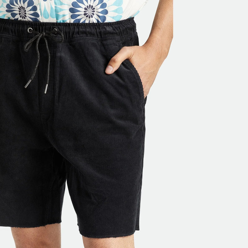 Load image into Gallery viewer, Close-up of a person wearing black corduroy shorts with a drawstring waist and side pockets, paired with a white top featuring a blue floral pattern, against a light gray background.
