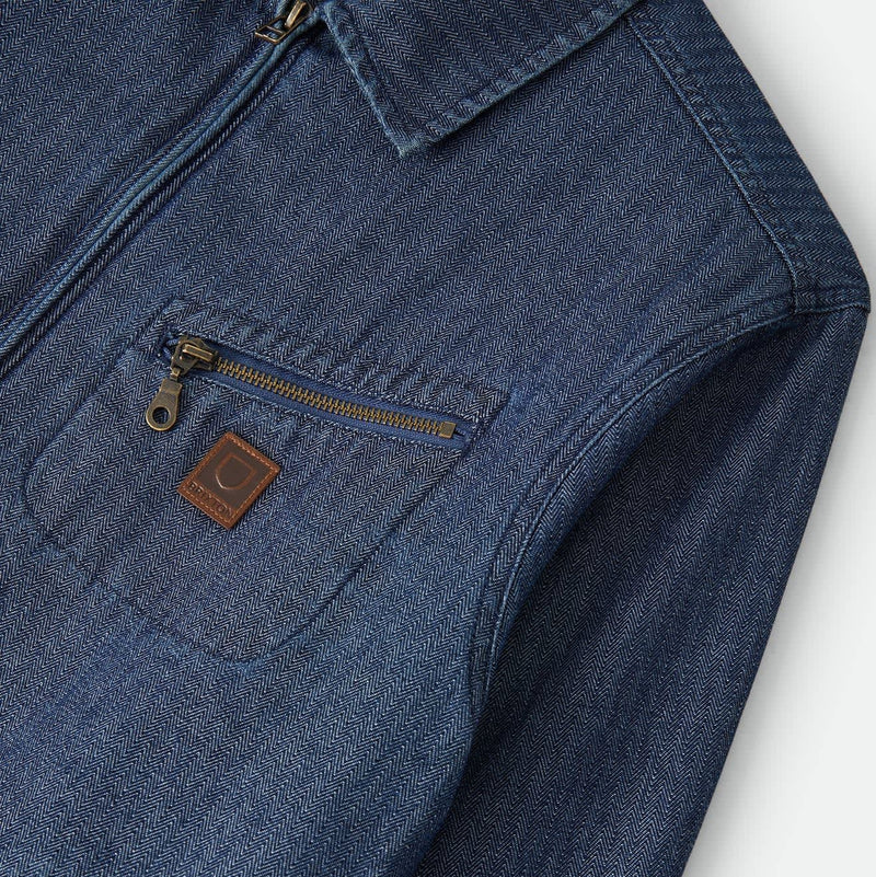 Load image into Gallery viewer, Close-up of a blue denim zip-front jacket or overshirt with a chest pocket and a small brown leather brand patch, shown against a light neutral background.
