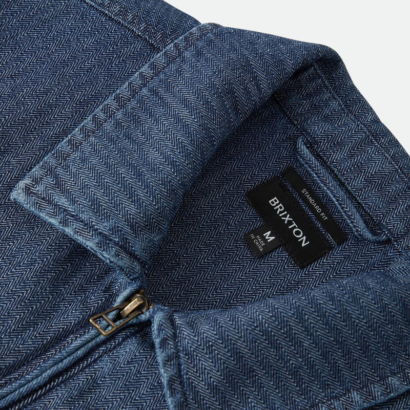 Load image into Gallery viewer, Close-up of a blue denim jacket showing the collar, a black BRIXTON brand label, and a small gold zipper pull against a light gray background.
