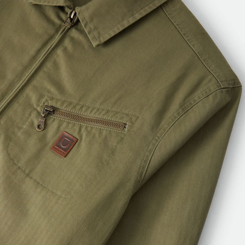 Load image into Gallery viewer, Close-up of an olive green jacket or overshirt showing a collar, front zipper, chest zip pocket, and a small brown brand patch on the pocket.
