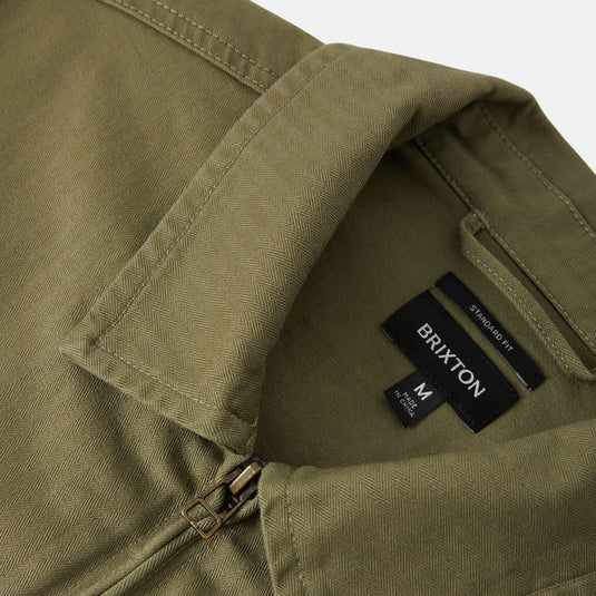 Close-up of an olive green collared shirt with a zipper at the collar, showing a black neck label that reads BRIXTON.
