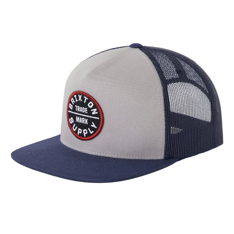 Load image into Gallery viewer, A Brixton trucker hat featuring a light gray front panel, navy blue flat brim, and navy blue mesh back. The front panel has a circular embroidered patch with the brand name.
