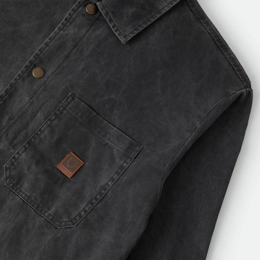Close-up of a dark charcoal gray waxed cotton shirt jacket with a chest pocket and a small brown leather brand patch.