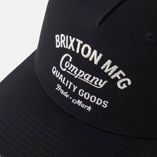 A close-up of a black baseball cap featuring white embroidered text that reads 'BRIXTON MFG Company QUALITY GOODS Trade Mark' on a light gray background.