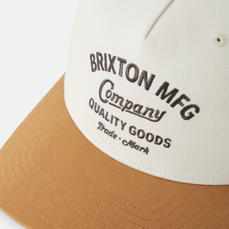 Load image into Gallery viewer, A close-up of a baseball cap with an off-white crown and a tan curved brim, featuring black embroidered text that reads 'BRIXTON MFG Company Quality Goods Trade Mark'.
