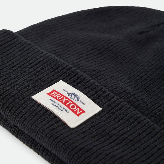 Close-up of a black ribbed knit beanie with a white Brixton brand patch on the cuff, photographed on a light gray background.