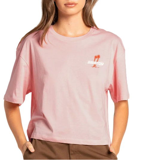 A woman wearing a light pink short-sleeve t-shirt with a small orange and white graphic on the left chest, paired with brown pants, against a plain white background.