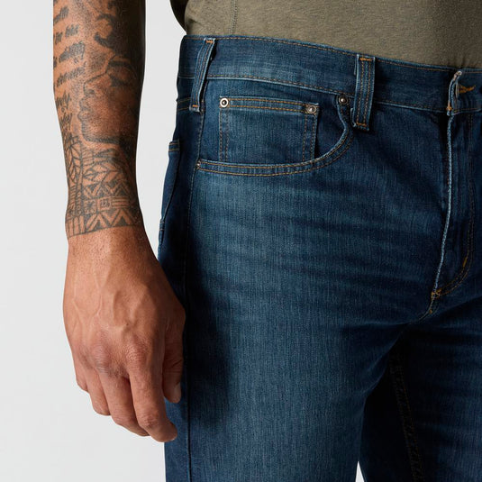 Close-up of a person wearing blue jeans with a tattooed arm on a plain background