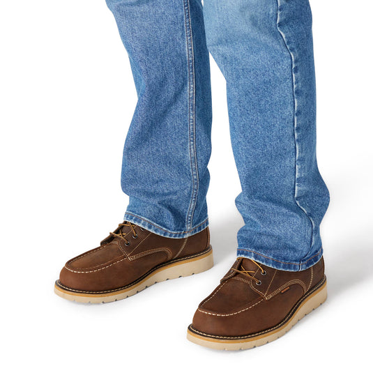 A person wearing blue jeans and brown suede lace-up shoes with light tan soles, shown from the calves down on a white studio background.