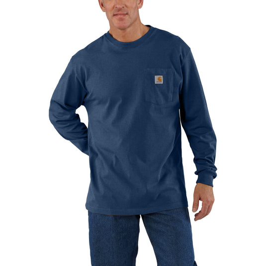 A man wearing a dark blue long-sleeve pocket t-shirt with a small Carhartt logo on the chest pocket, paired with dark blue jeans, against a plain white background.