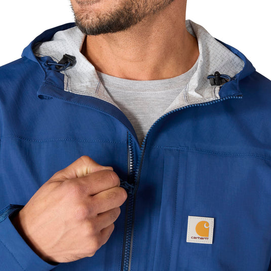 A close-up of a man wearing a blue Carhartt hooded jacket over a gray t-shirt, with his hand adjusting the zipper. The Carhartt logo patch is visible on the left chest.