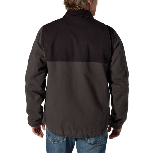 A man viewed from the back wearing a dark color-blocked jacket with a black upper back and shoulders and a charcoal gray lower back, paired with blue jeans, on a white background.