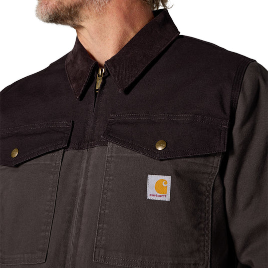 Close-up of a person wearing a Carhartt work jacket, showing the collar, zipper, chest pockets, and a visible Carhartt logo patch on a dark brown and charcoal fabric.