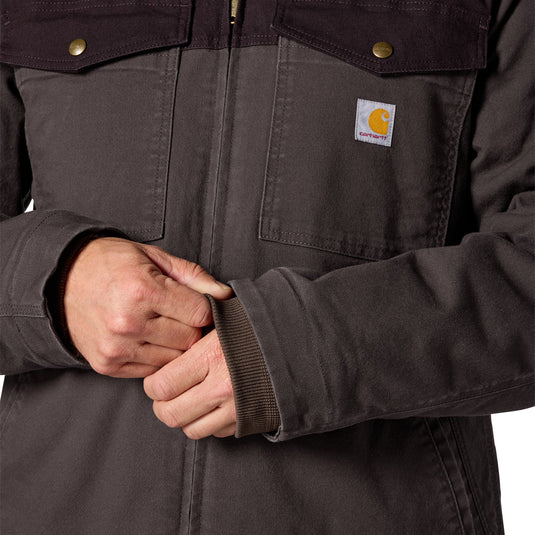 Close-up of a person wearing a dark brown Carhartt work jacket, showing the chest pocket, brass snap buttons, and the signature Carhartt logo patch on the pocket.