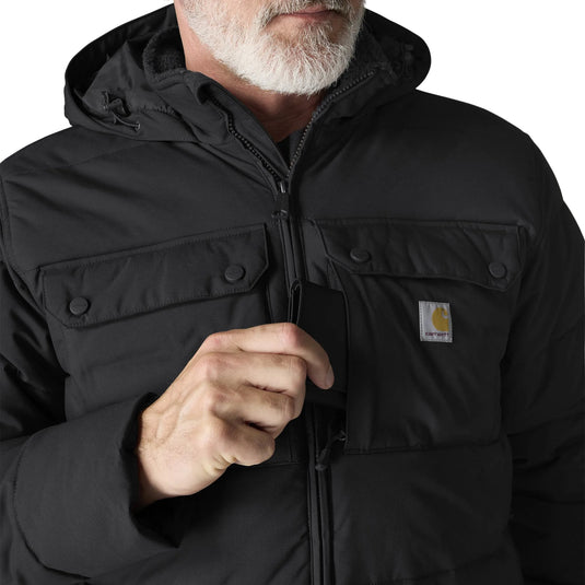 A close-up of a man wearing a black hooded puffer jacket with a visible Carhartt logo patch on the chest pocket, against a light gray background.