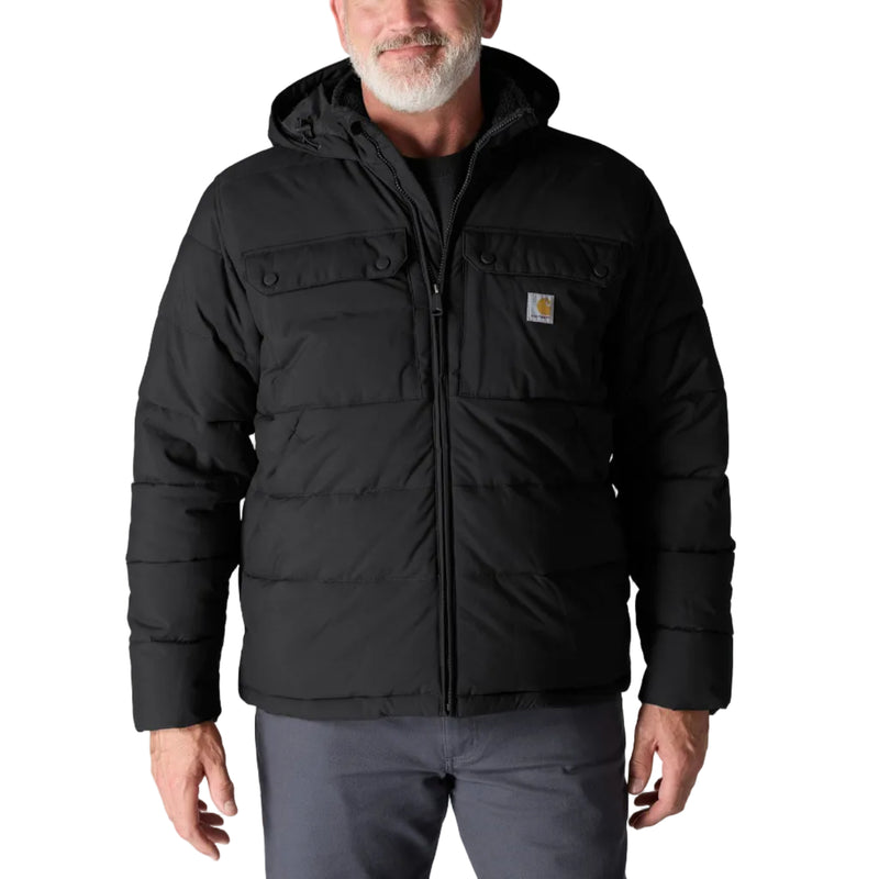 Load image into Gallery viewer, A man wearing a black hooded puffer jacket with a full front zipper and a small Carhartt logo patch on the chest, paired with gray pants, against a light gray studio background.
