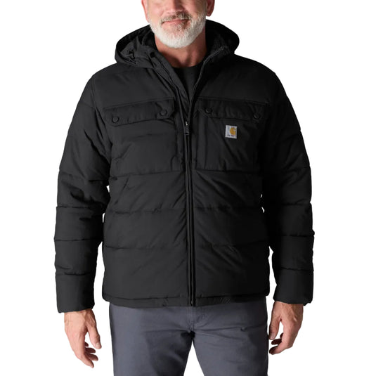 A man wearing a black hooded puffer jacket with a full front zipper and a small Carhartt logo patch on the chest, paired with gray pants, against a light gray studio background.