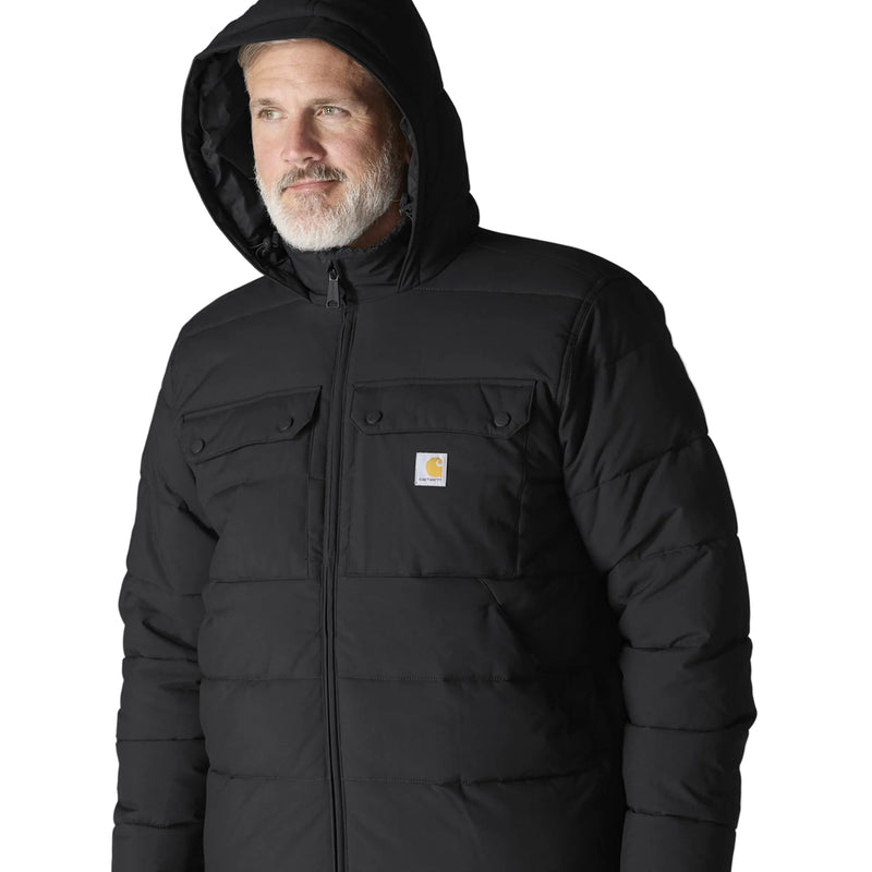 Load image into Gallery viewer, A man wearing a black hooded puffer jacket with a visible Carhartt logo patch on the chest, photographed against a light gray studio background.
