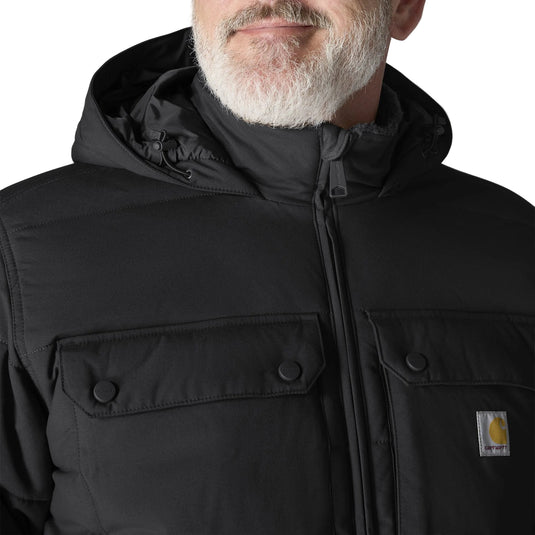Close-up of a man wearing a black hooded puffer jacket with a high collar, front zipper, and two flap chest pockets. A small Carhartt logo patch is visible on the left chest.