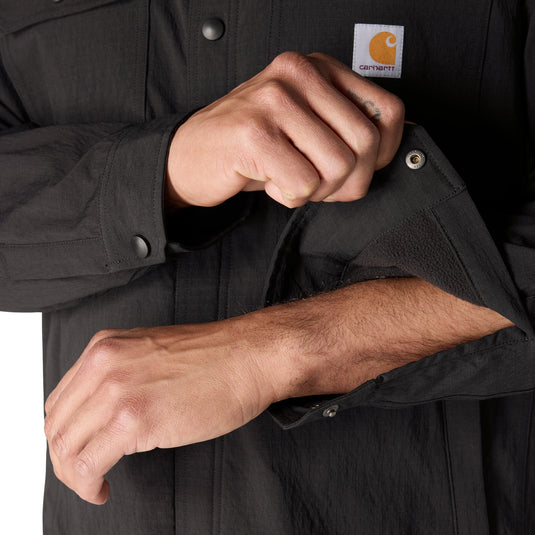 Close-up of a person wearing a black Carhartt work shirt, pulling up the sleeve to show the cuff and button detail. The Carhartt logo patch is visible on the chest.