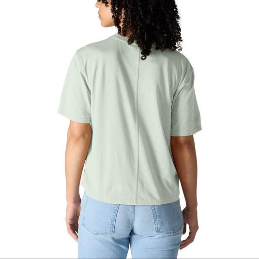 A woman with curly black hair is shown from the back wearing a light sage green short-sleeve t-shirt and light blue jeans against a solid white background.
