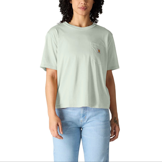 A woman with curly black hair wearing a light sage green short-sleeve pocket t-shirt and light blue jeans, standing against a solid white background.
