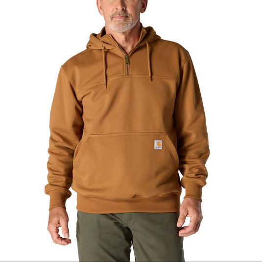 A man wearing a brown pullover hoodie with a half-zip neckline, drawstrings, and a kangaroo pocket, paired with dark green pants, against a plain white background.