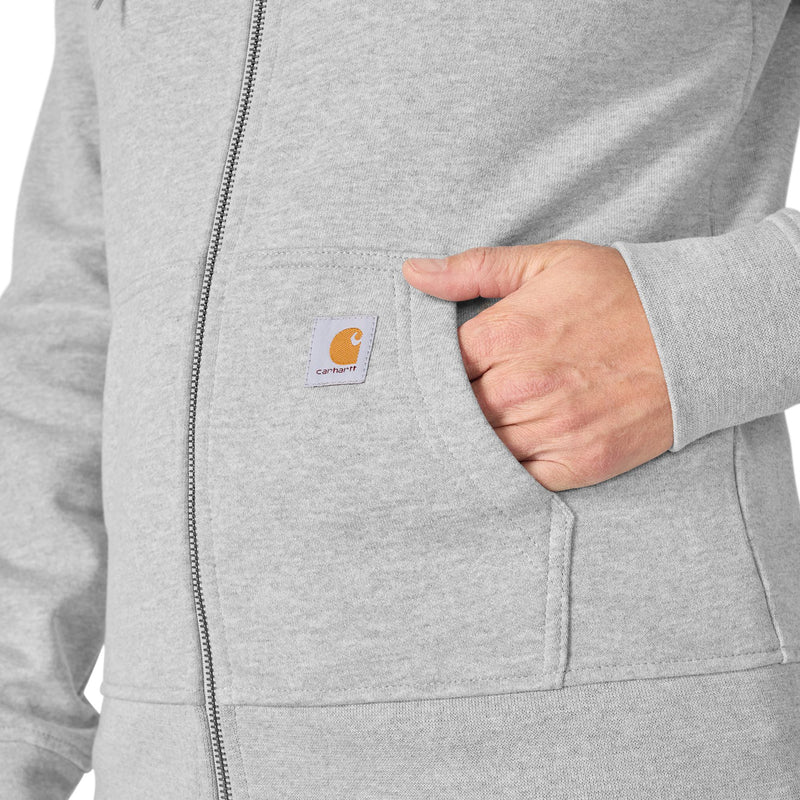 Load image into Gallery viewer, Close-up of a person wearing a light heather gray zip-up hoodie, with a hand in the side pocket showing the Carhartt logo patch.
