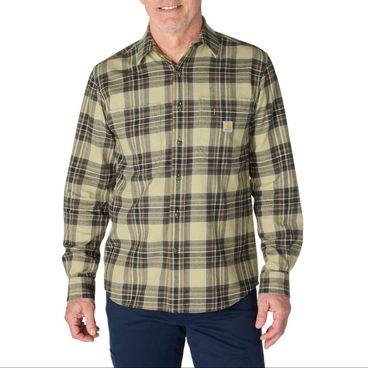 A man wearing a Carhartt long-sleeve plaid button-down shirt in olive green and beige with dark brown and black lines, paired with navy pants, against a plain white background.