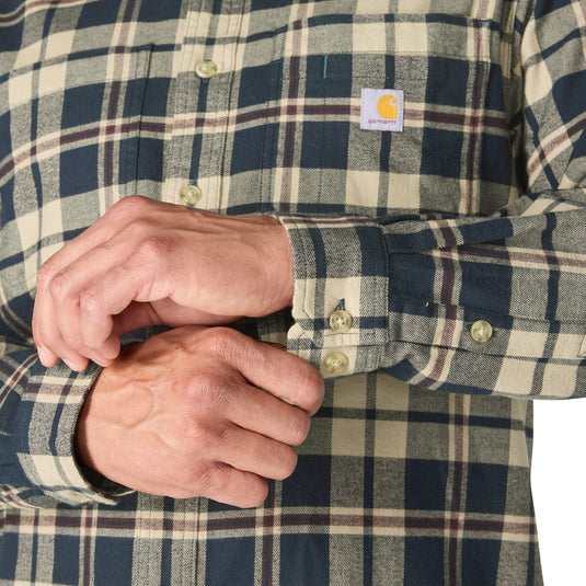 Close-up of a person wearing a Carhartt plaid button-up shirt, adjusting the cuff. The shirt features a navy, beige, and brown plaid pattern with a visible Carhartt logo patch on the chest.