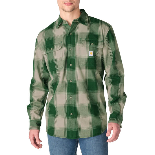 A man wearing a green and beige plaid Carhartt flannel shirt with blue jeans, standing against a plain white background.