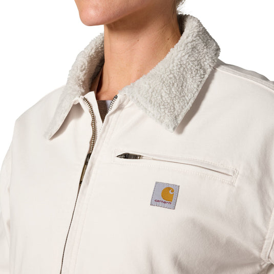 Close-up of a person wearing a Carhartt work jacket with a cream-colored sherpa collar and a zippered chest pocket. The Carhartt brand patch is visible on the chest pocket.