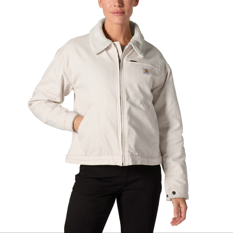 Load image into Gallery viewer, A woman modeling a light beige Carhartt jacket with a sherpa collar, front zipper, and chest pocket, paired with black pants against a plain white background.
