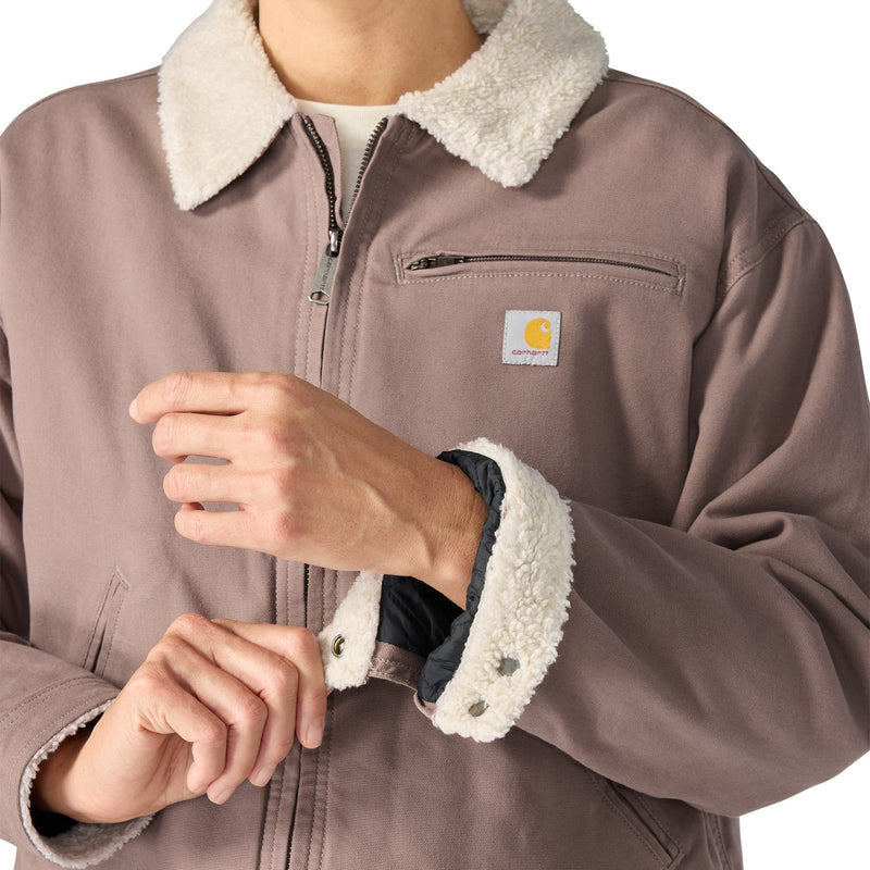 Load image into Gallery viewer, A close-up of a person wearing a brown Carhartt jacket with a cream sherpa collar and matching sherpa-lined cuffs, showing the brand patch on the chest pocket.
