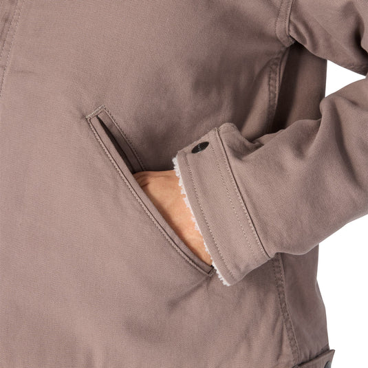 Close-up of a taupe-brown utility jacket with a side pocket, showing a hand reaching into the pocket and the interior fleece lining.