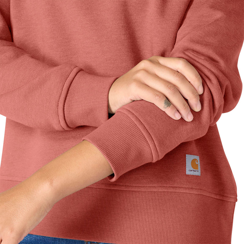 Load image into Gallery viewer, A close-up of a person wearing a dusty rose long-sleeve sweatshirt, showing the ribbed cuffs, a small Carhartt logo patch on the lower front, and blue jeans at the bottom edge.
